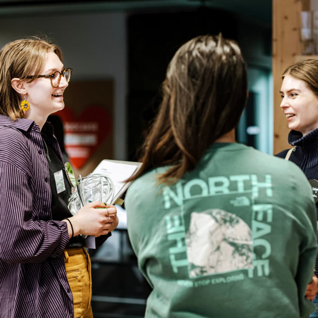 Bilden visar tre personer som står och pratar med varandra i ett inomhusutrymme som ser ut att vara en kontorsmiljö eller ett mötesrum. Personen till vänster i bilden, som har glasögon och en randig skjorta, håller i ett glas och ser ut att vara i en livlig konversation med de andra två. Personen i mitten har ryggen mot kameran och bär en grön tröja med texten "NORTH PEACE" och en bild på en bergstopp. Personen till höger i bilden, som är delvis vänd mot kameran, har en mörkblå tröja och ser ut att lyssna uppmärksamt på vad den första personen säger. Bakgrunden är suddig men man kan urskilja en vattenkylare och några glas på en hylla. Atmosfären verkar avslappnad och vänlig.